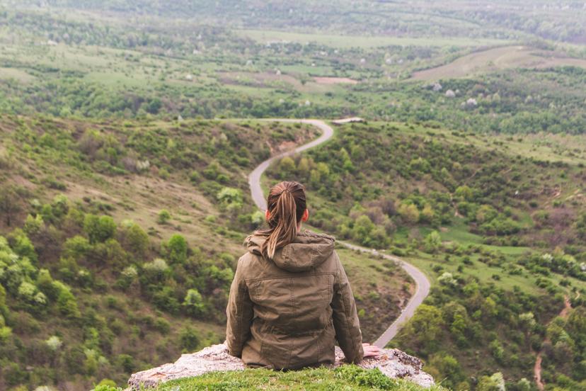A person in a hooded jacket sits on a rocky outcrop, overlooking a lush, forested valley with winding roads below.