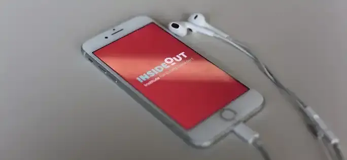 A white smartphone with a red "Inside Out" logo displayed on the screen, resting on a gray surface and connected to a pair of white earphones.