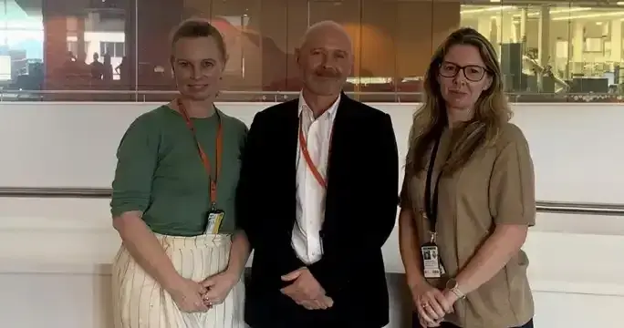 Three individuals, two women and one man, are standing together in what appears to be a professional or office setting, with a glass wall or window visible in the background.