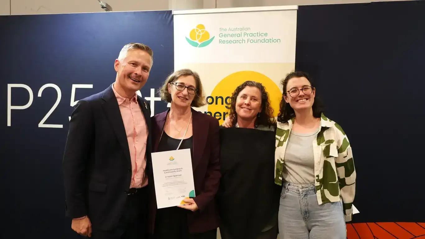The image shows four individuals standing in front of a backdrop with the logo of the General Practice Research Foundation. The individuals appear to be posing for a photograph, with one holding a certificate or award.