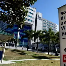 The image shows a modern, multi-story building surrounded by palm trees and a grassy area in the foreground.