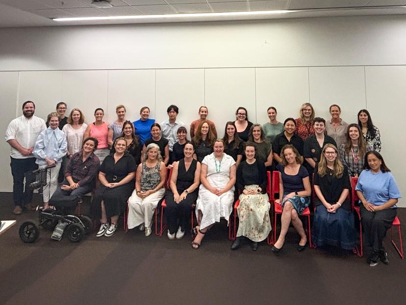 A large group of people, both men and women, are seated and standing together in a room with white walls and a tiled ceiling.