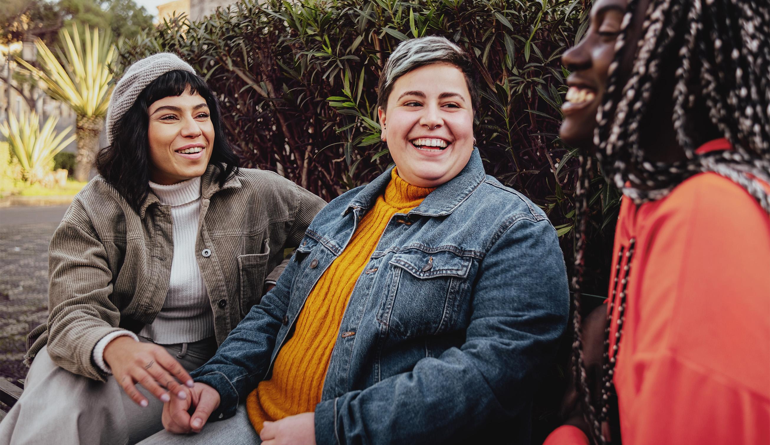 Three friends laugh together outdoors, with lush green foliage forming a soft background.