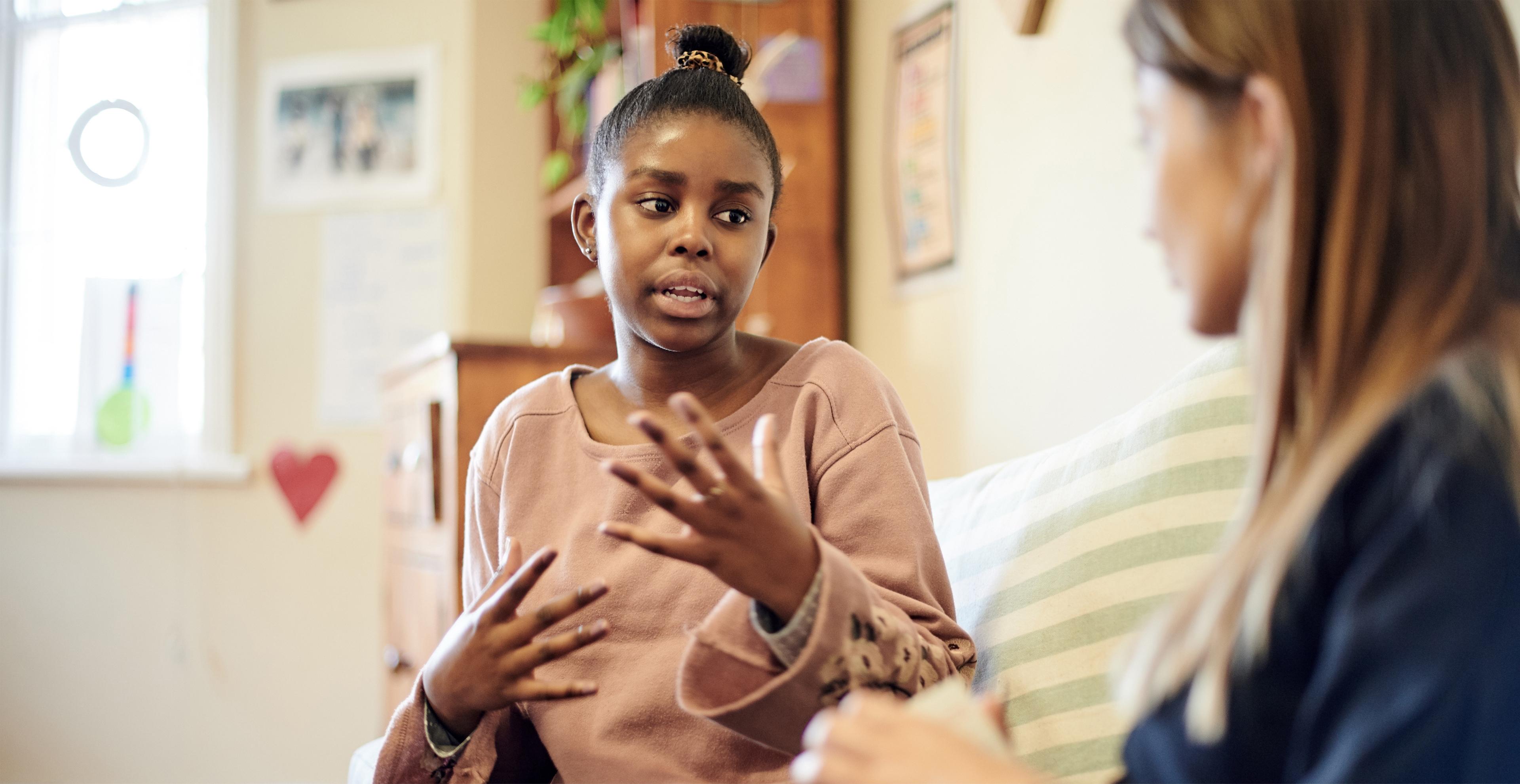 A young Black woman gestures with her hands while speaking to another woman in a brightly lit room with a wooden dresser and framed pictures in the background.