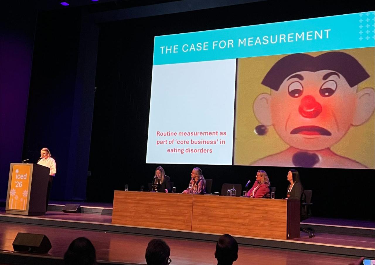 A speaker stands at a podium on a stage in front of a large screen displaying "THE CASE FOR MEASUREMENT" and a cartoonish face, while five panelists sit at a table below.