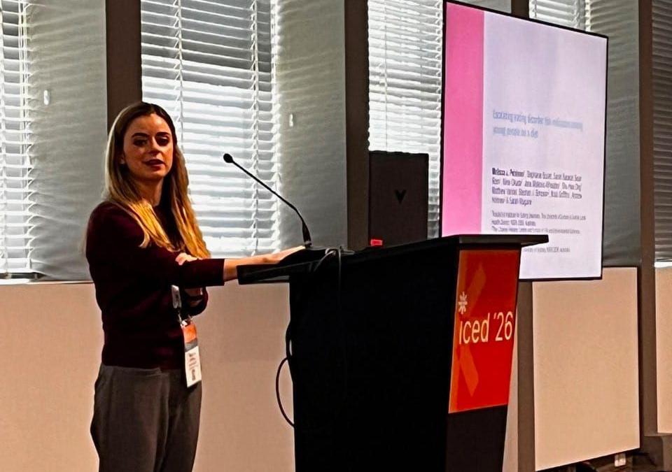 A woman stands at a podium in front of a presentation screen, with blinds covering a window in the background.