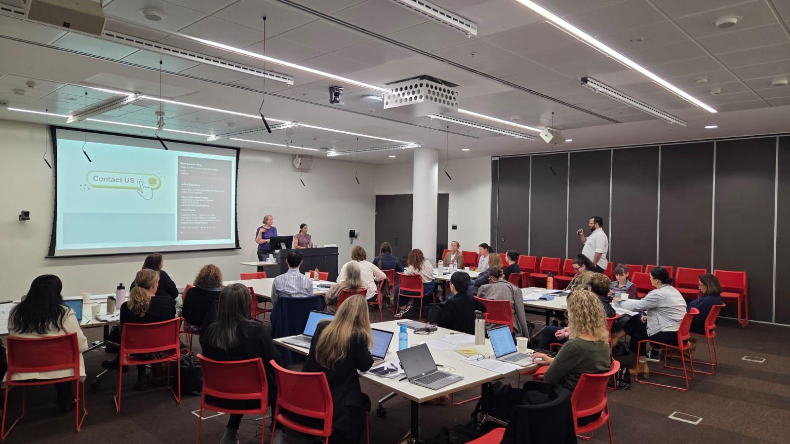In the foreground, people sit at tables with laptops, while in the background, a presentation is projected onto a screen.